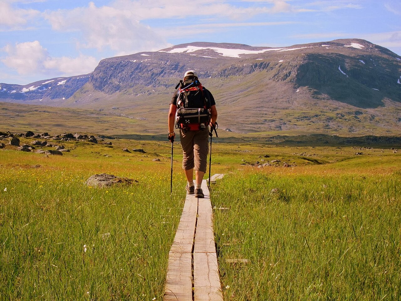 Hiker with poles and a full pack walking a wooden boardwalk across open fjäll, mountains ahead.