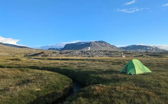 Green tent pitched beside a small stream on open tundra, mountains behind.